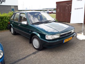 Een groene vierdeurs hatchback staat geparkeerd op een verhard terrein nabij een houten hek en een gebouw, voorzien van een geel kenteken met de code "SG-45-ZG". Dit voertuig van Elk Merk Waardig valt op door zijn levendige kleur.