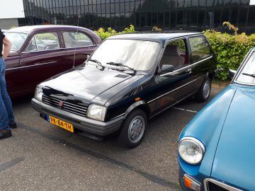 Een zwarte klassieke compacte auto van Elk Merk Waardig staat buiten geparkeerd tussen twee andere voertuigen.