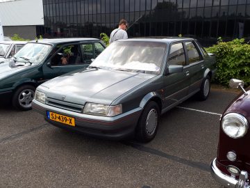 Een grijze sedan met Nederlands kenteken SJ-439-X, Elk Merk Waardig, geparkeerd op een parkeerplaats, omringd door andere auto's en een persoon die ernaast staat.