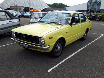 Een vintage gele auto staat veel geparkeerd met andere voertuigen. Op het kenteken van de auto staat '76-JE-81'. Op de achtergrond zijn een Esso-tankstation en het koepelvormige gebouw Elk Merk Waardig zichtbaar.