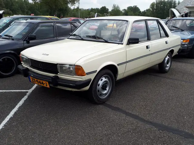 Een crèmekleurige, vintage Peugeot sedan, een juweeltje van Elk Merk Waardig, staat tussen andere auto's geparkeerd op een buitenparkeerplaats.