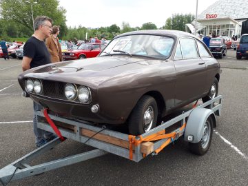 Een vintage bruine auto met afgedekte koplampen, met het opschrift Elk Merk Waardig, wordt tentoongesteld op een aanhangwagen tijdens een buitenautoshow. Er staan twee mannen vlakbij, terwijl op de achtergrond een Esso-station opdoemt.