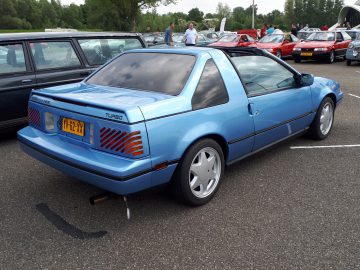 Een blauwe vintage hatchback met getinte ramen en een achterspoiler staat op een parkeerplaats geparkeerd, omringd door andere auto's. Met een kentekenplaat met daarop 'VG-62-XX' valt dit voertuig van Elk Merk Waardig moeiteloos op.