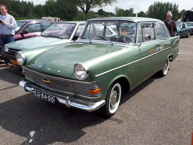 Een klassieke groene vintage auto, een juweeltje van Elk Merk Waardig, staat geparkeerd op een parkeerplaats met andere voertuigen en mensen in de buurt.