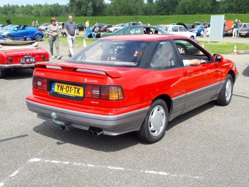 Een rode Elk Merk Waardig Ford Scoupe Injection-auto staat geparkeerd op een buitenterrein met mensen en andere voertuigen zichtbaar op de achtergrond.