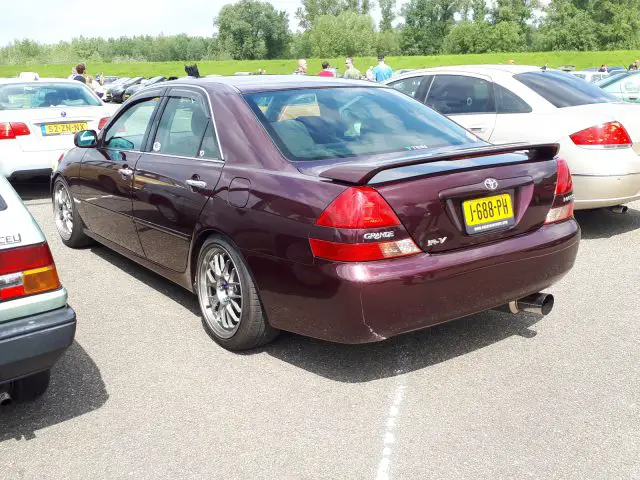 Een paarse Toyota Mark II sedan, die er nogal Elk Merk Waardig uitziet, staat op een zonnige dag tussen andere auto's geparkeerd op een parkeerplaats. De auto is voorzien van een spoiler en een aftermarket uitlaat. Op het kenteken staat "1-688-PA." Op de achtergrond zijn bomen zichtbaar.