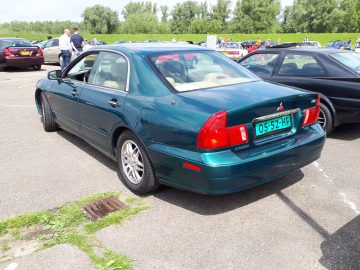 Een groene Mitsubishi Galant, geparkeerd op een parkeerplaats met andere voertuigen zichtbaar op de achtergrond, valt op als Elk Merk Waardig. Twee mensen staan bij de auto en achter de parkeerplaats is een grasveld te zien.