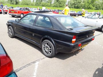 Tussen verschillende voertuigen staat een strakke zwarte sportcoupé, versierd met een sticker van Elk Merk Waardig op de achterruit, geparkeerd. De opvallende achterspoiler valt op, terwijl verschillende mensen op de achtergrond bijdragen aan de bruisende sfeer van de scène.