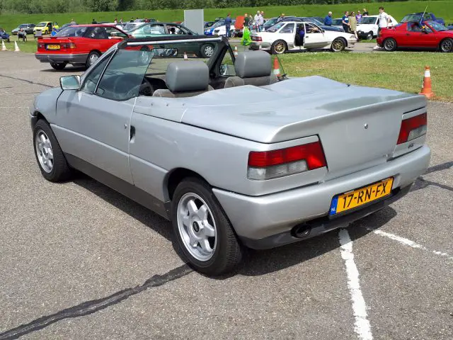 Een zilveren cabriolet, heel Elk Merk Waardig, staat geparkeerd op een buitenevenement met andere auto's op de achtergrond. Op het kenteken staat "17-RN-FX.