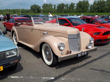 Een beige klassieke cabriolet van Elk Merk Waardig staat geparkeerd op een autoshow, omringd door andere voertuigen. Hij heeft een kentekenplaat met de tekst "DE-73-51" en heeft een strak, vintage ontwerp.