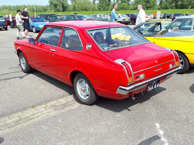 Een rode klassieke auto, versierd met het kenteken "NA 84", staat geparkeerd op een parkeerplaats omringd door andere vintage voertuigen. In de buurt staan mensen te praten en bewonderen de tentoongestelde collectie Elk Merk Waardig.