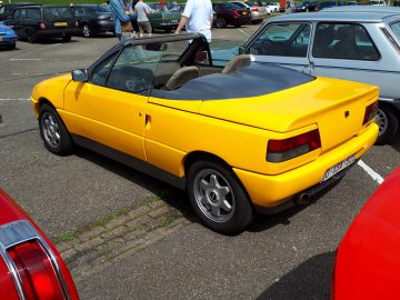 Een gele cabriolet, met trots het embleem van Elk Merk Waardig, staat geparkeerd op een parkeerplaats omringd door andere voertuigen. Met de bovenkant naar beneden zijn het Europese kenteken en twee voorstoelen zichtbaar.