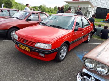 Een rode Peugeot sedan met Nederlands kenteken, Elk Merk Waardig, staat tussen andere klassieke auto's geparkeerd op een buitenevenement. Op de achtergrond zijn meerdere mensen zichtbaar.