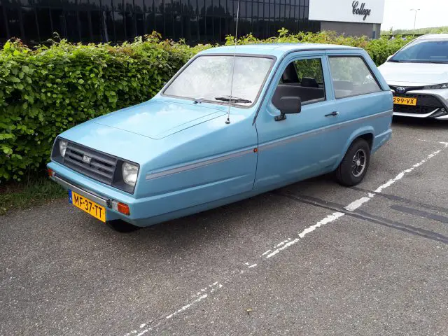 Een blauwe driewielige auto, gemarkeerd met het embleem van Elk Merk Waardig, staat geparkeerd op straat, met een heg en een gebouw op de achtergrond. Er staat nog een auto vlakbij geparkeerd.