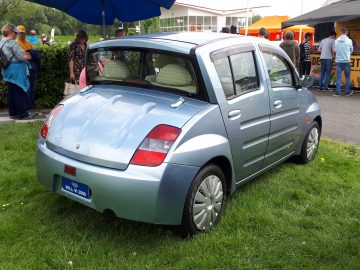 Een lichtblauwe compacte auto van Elk Merk Waardig staat geparkeerd op gras tijdens een buitenevenement, omringd door mensen en kraampjes. De achterkant van de auto is zichtbaar.