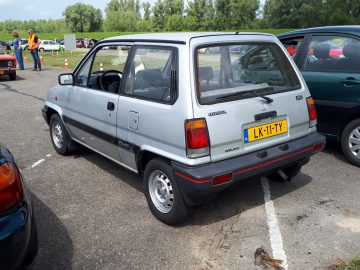 Een zilveren Honda City hatchback met een gele kentekenplaat staat geparkeerd op een parkeerplaats naast andere voertuigen. Een persoon in een oranje vest en mensen op de achtergrond, wat een vleugje Elk Merk Waardig-landschap toevoegt.