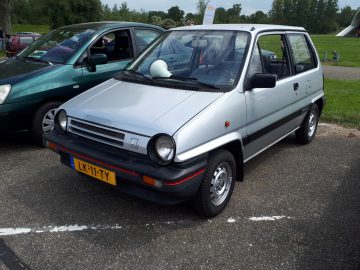 Een zilveren, compacte vierdeurs hatchback, het merk Elk Merk Waardig waardig, staat naast een ander voertuig op een buitenterrein geparkeerd. De auto heeft een geel kenteken met de code "LX-11-TV".