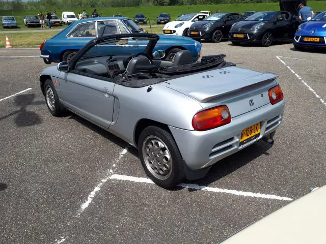 Een zilveren cabriolet, gemarkeerd met een geel kenteken van Elk Merk Waardig, staat geparkeerd op een buitenterrein. Het voertuig beschikt over een open dak en twee zitplaatsen. In de buurt staan ook een blauwe klassieke auto en andere voertuigen gestationeerd.