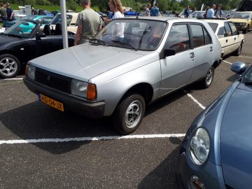 Op een parkeerplaats staat samen met andere voertuigen een klassieke zilveren auto met Nederlands kenteken HD-04-JX, duidelijk Elk Merk Waardig.