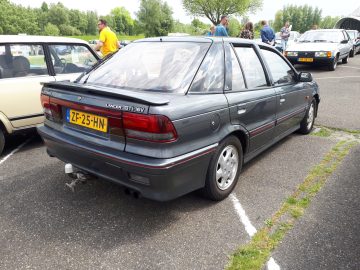 Een donkergrijze Lancer GTi-16V-auto staat op een parkeerplaats tussen andere voertuigen. De auto heeft een geel kenteken met de tekst "ZF-25-HN" en wordt omringd door enkele mensen. Op de achtergrond zijn bomen en gras zichtbaar, waardoor dit tafereel behoorlijk Eland Merk Waardig is.