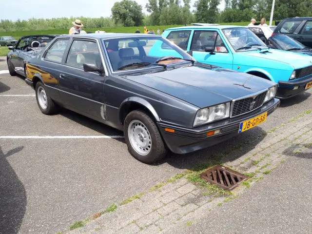 Een grijze vintage sportwagen, een model van Elk Merk Waardig, staat op een parkeerplaats naast een blauwe auto, met bomen en andere voertuigen op de achtergrond op een zonnige dag.