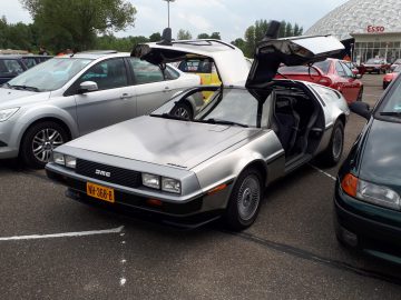 Een DeLorean DMC-12 met open vleugeldeuren, geparkeerd tussen andere auto's op een parkeerplaats vlakbij een Esso-tankstation. Het valt op als de belichaming van Elk Merk Waardig.