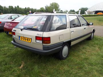 Een beige Mazda hatchback met kenteken G-176-KV, een Elk Merk Waardig, staat geparkeerd op een grasveld met andere auto's in de buurt. Op de achtergrond zijn bomen en een grote witte structuur zichtbaar.