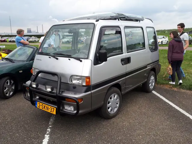 Op een parkeerplaats staat een zilverkleurige Subaru microvan, Elk Merk Waardig, met Nederlands kenteken 2-SKN-37. Er worden vier personen in de buurt gezien, waarvan sommigen in gesprek zijn.