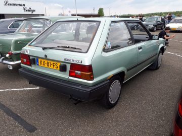 Een lichtgroene Toyota Tercel hatchback staat samen met andere auto's geparkeerd, met een geel kenteken met de tekst "KX-81-FS". Dit voertuig is een echt voorbeeld van de uitdrukking "Elk Merk Waardig.