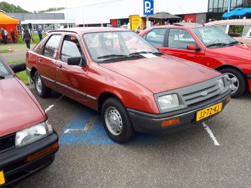 Een rode Ford sedan, versierd met een geel kenteken "JJ-72-XJ", staat geparkeerd op een buitenterrein. Vlakbij komt nog een rode auto in beeld, terwijl op de achtergrond een wit gebouw hoog staat: echt Elk Merk Waardig.