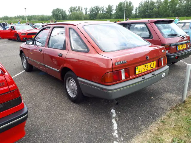 Een rode Ford Sierra, die er echt Merk Waardig uitziet, staat geparkeerd op een buitenterrein tussen andere voertuigen, met uitzicht vanaf de bestuurderszijde aan de achterkant.