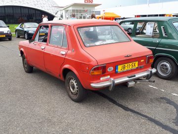 Een vintage rode sedan met een Nederlands kenteken, waarop trots 'Elk Merk Waardig' staat, staat geparkeerd op een autoshow naast andere klassieke voertuigen, waaronder een nabijgelegen groene auto. Op de achtergrond staan een Esso-tankstation en evenementententen.