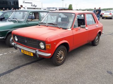 Op een parkeerplaats staat een rode vintage vierdeurs auto met zwarte bekleding, voorzien van kenteken J8-11-SX. Op de achtergrond zijn andere klassieke auto's en een gebouw met het opschrift "Louwman College" zichtbaar, wat een sfeer creëert waarin elk voertuig Elk Merk Waardig is.