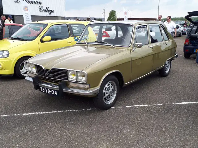 Een vintage olijfgroene auto, echt Elk Merk Waardig, staat geparkeerd op een asfaltoppervlak, geflankeerd door andere kleurrijke auto's. Op het kenteken staat "26-YB-04.