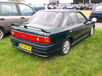 Een groene Mazda Familia sedan met Nederlands kenteken HX-GH-34, voorzien van de Elk Merk Waardig-norm, staat op een grasveld bij een autobijeenkomst geparkeerd, naast andere voertuigen.