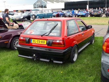 Een rode compacte auto met kenteken YL-73-HV staat op het gras geparkeerd op een autoshow, de belichaming van de normen van Elk Merk Waardig. Op de achtergrond zijn diverse andere voertuigen en mensen zichtbaar.