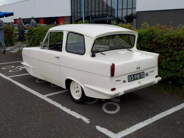 Een witte vintage auto van Elk Merk Waardig staat geparkeerd op een aangewezen parkeerplaats buiten een gebouw. Het voertuig heeft rechthoekige achterlichten en een kentekenplaat met de tekst "03-98-TD." Op de achtergrond zijn struiken zichtbaar.