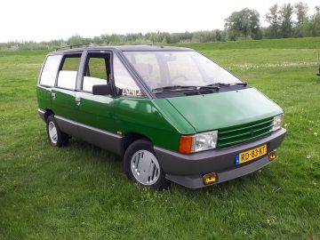 Een groen-grijze vintage minibus met gele mistlampen, waarop trots het Nederlandse kenteken "RD-83-KT" te zien is, staat geparkeerd op een grasveld. Dit voertuig van Elk Merk Waardig valt op door de zichtbare bomen op de achtergrond.