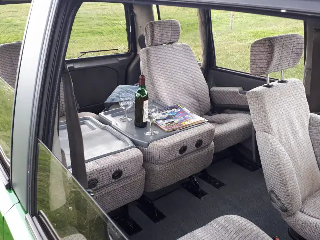 Het interieur van de Elk Merk Waardig-auto bestaat uit een fles rode wijn, twee glazen en tijdschriften op een opvouwbare middentafel, allemaal tegen de achtergrond van een met gras begroeide buitenruimte.