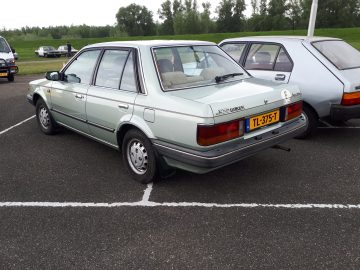 Een zilveren Mazda 323 GLX sedan, geparkeerd op een parkeerplaats naast een lichtblauwe auto, heeft het kenteken "TL-375-T". Op de achtergrond zijn bomen en gras zichtbaar, waardoor dit tafereel echt Elanden Merk Waardig is.