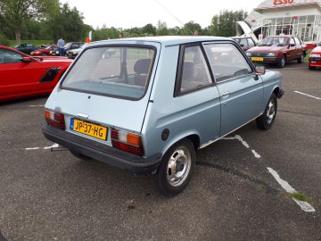 Een lichtblauwe hatchback staat op een parkeerplaats samen met andere voertuigen onder toeziend oog van een bord Elk Merk Waardig. De auto heeft een geel kenteken met de tekst "JP-37-HG." Op de achtergrond is er een Esso-tankstation.