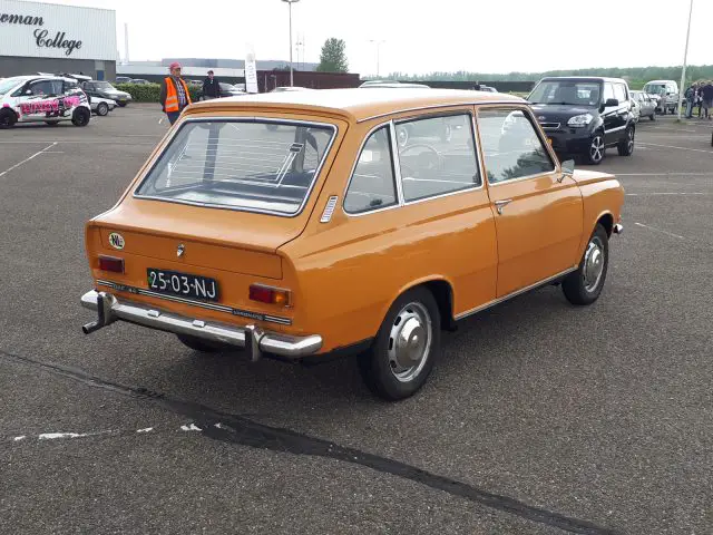 Een vintage oranje auto geparkeerd in een openluchttentoonstellingsruimte met andere voertuigen en een universiteitsgebouw op de achtergrond, dat de charme van Elk Merk Waardig laat zien.