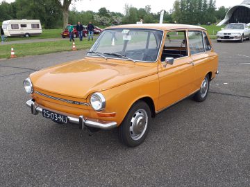 Een oranje klassieke auto van Elk Merk Waardig, geparkeerd op een parkeerplaats buiten, heeft een vintage kentekenplaat "25-03-NJ" op de voorbumper. Op de achtergrond zijn andere auto's en enkele mensen zichtbaar.