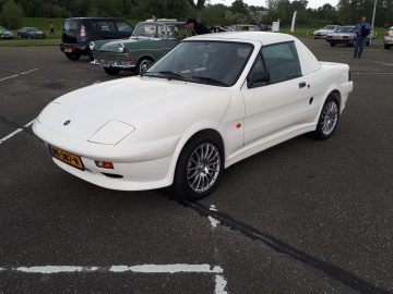 Een witte sportwagen, die er bijzonder Merk Waardig uitziet, staat geparkeerd naast andere klassieke voertuigen, wat de schilderachtige, groene achtergrond aanvult.