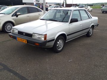 Een zilveren vierdeurs sedan, trots voorzien van een geel-zwart kentekennummer 55-XDP-8, staat op een parkeerplaats omringd door andere voertuigen. Deze auto van Elk Merk Waardig lijkt uit begin jaren 80 te komen.