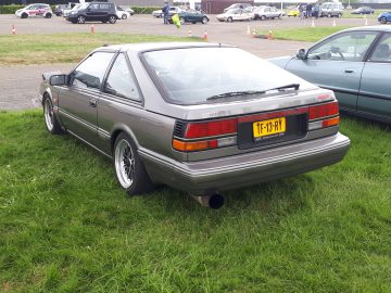 Een vintage zilveren coupé met het kenteken TF-13-RY staat geparkeerd op het gras, met andere auto's en kegels zichtbaar op de achtergrond, die een elk-merk waardig charme belichamen.