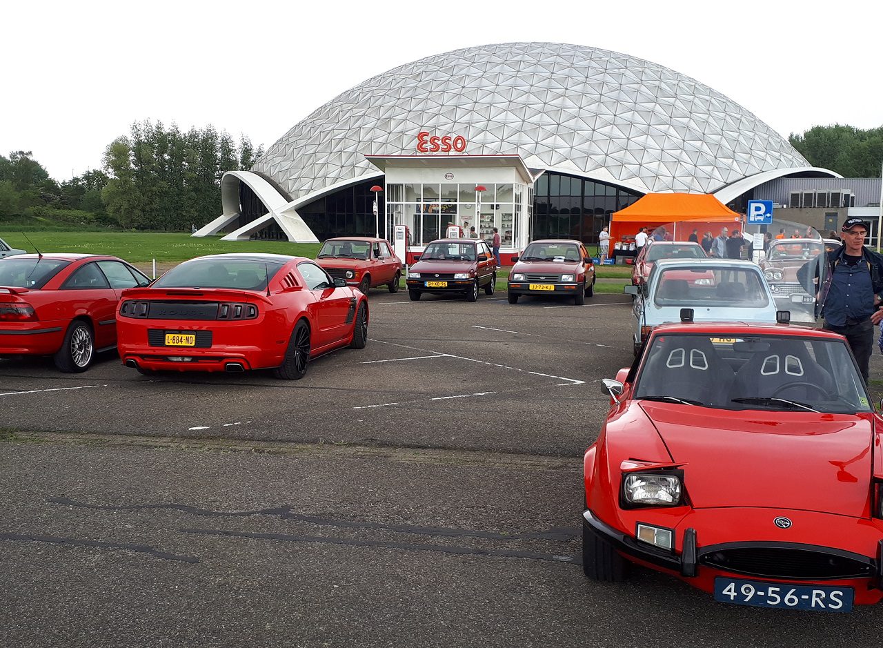 Een groep oldtimers, overwegend rood, staat geparkeerd voor een Esso-station met een geodetische koepelstructuur. Verschillende mensen, getroffen door de Elk Merk Waardig-charme van het tafereel, staan bij de auto's.