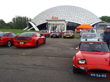 Een groep oldtimers, overwegend rood, staat geparkeerd voor een Esso-station met een geodetische koepelstructuur. Verschillende mensen, getroffen door de Elk Merk Waardig-charme van het tafereel, staan bij de auto's.