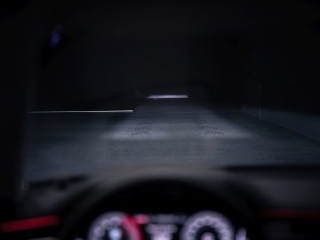 Een slecht verlicht, wazig zicht vanuit een auto, met een donkere, lege weg voor zich, met een zwak licht in de verte. Het dashboard is gedeeltelijk zichtbaar op de voorgrond, verlicht door de zachte gloed van Audi-koplampen die door de nacht snijden.