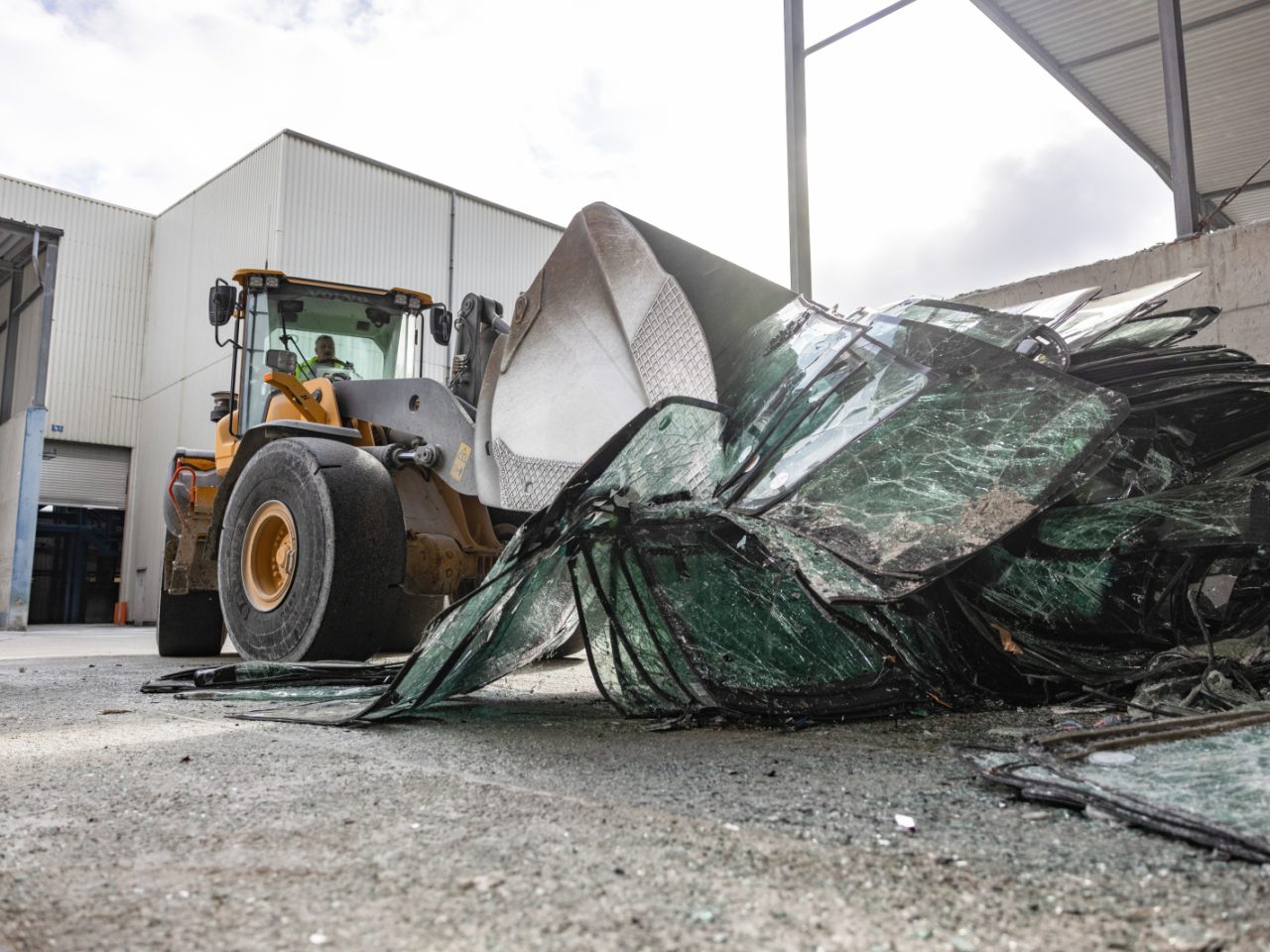 We zien een front-end-lader gebroken glasstukken opscheppen in een industriële omgeving met magazijnen op de achtergrond, terwijl een Audi Q4 e-tron voorbij komt, die een mix van ruige industrie en gestroomlijnde auto-innovatie laat zien.